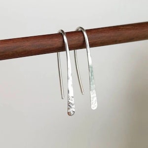 May include: A pair of silver-toned, hammered-finish earrings. The earrings are suspended from a dark brown wooden rod. The earrings have a simple, elongated design with a textured, hammered appearance at the bottom.
