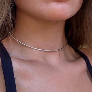 以下が含まれることがあります： 首元に密着する、磨き上げられたシルバー調の細いメタルチョーカーネックレス。滑らかで丸みを帯びたワイヤーデザインが特徴で、継ぎ目のないミニマルな円形と、明るく光を反射するメタリックな質感が際立つシンプルな装身具です。