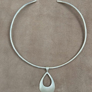 以下が含まれることがあります： 涙型のペンダントが付いたシルバーカラーのネックレス。 ネックレスは首の周りにフィットする硬い円形デザインです。 ペンダントは、切り抜かれた中央部を備えた、滑らかで研磨された涙の形をしています。 背景は無地のニュートラルブラウンです。