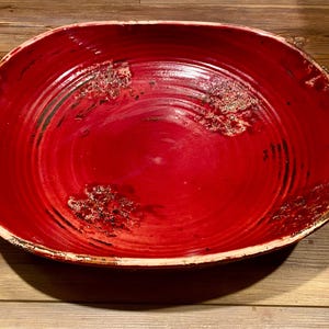 May include: A vibrant red ceramic serving bowl with a rustic, textured finish. The bowl has a wide, oval shape with twisted rope-like handles on each side. The interior shows concentric circles.