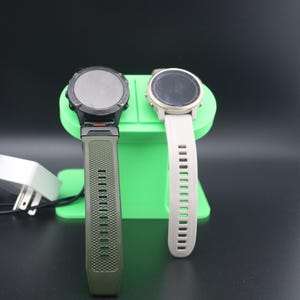 May include: Two smartwatches with circular faces and straps, one olive green and one off-white, are on a green stand. A black charging cable and power adapter are in the foreground. The watches appear to be fitness trackers.