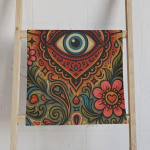 Puede incluir: Una toalla colorida con un diseño de ojo, flores y patrones de cachemira. La toalla presenta un ojo grande en el centro con un iris azul, rodeado de detalles rojos, naranjas y amarillos. Elementos florales y de cachemira están presentes.