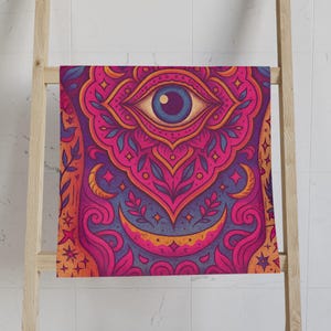 Puede incluir: Una toalla colorida con un diseño detallado de ojo. La toalla presenta un ojo central con iris azul, rodeado de intrincados patrones en rosa, morado, naranja y amarillo. El diseño incluye lunas crecientes, estrellas y elementos florales.