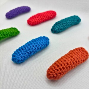 May include: A collection of small, oblong hair clips covered in vibrant, hand-crocheted yarn. The set includes individual clips in solid colors: purple, red, teal, light pink, bright green, royal blue, and orange, all featuring a textured stitch.