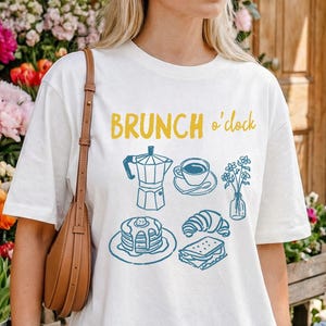 May include: White t-shirt with the text "BRUNCH o'clock" in yellow and blue. The shirt features illustrations of a coffee pot, cup, flowers, pancakes, croissant, and sandwich. The shirt is worn by a person.