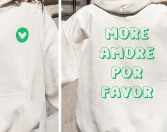 May include: Sand-colored hoodie with a front pocket and a green heart graphic. The back of the hoodie features the words "MORE AMORE POR FAVOR" in green, bubble-style letters. The word "sand" is printed on a circular label.