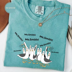 May include: Seafoam green Comfort Colors t-shirt with a graphic of seagulls yelling "Ms. Smith!". The shirt has a crew neck and short sleeves. A gold necklace with a pendant is draped across the shirt.