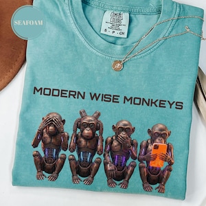May include: Seafoam green t-shirt with the text "MODERN WISE MONKEYS" and a graphic of four robotic monkeys. The monkeys are in the "see no evil, hear no evil, speak no evil" poses, with one holding a phone. A gold necklace is draped across the shirt.