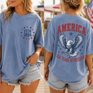 May include: A blue short-sleeve t-shirt with a vintage wash. The front features a circle of red and blue stars surrounding the text 1776 2026. The back displays a large bald eagle graphic with the text AMERICA 250 YEARS OF FREEDOM in red.