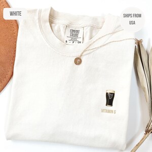 May include: Cream-colored t-shirt with a small graphic of a pint of stout and the text "VITAMIN G". A gold necklace with a round pendant rests on the shirt. The shirt is made of a soft material.