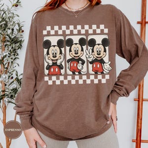 Puede incluir: Una camiseta marr&oacute;n de manga larga con un gr&aacute;fico de Mickey Mouse en tres poses diferentes. Las im&aacute;genes de Mickey Mouse est&aacute;n enmarcadas en cuadros blancos y negros. La camiseta tiene cuello redondo y un corte holgado.