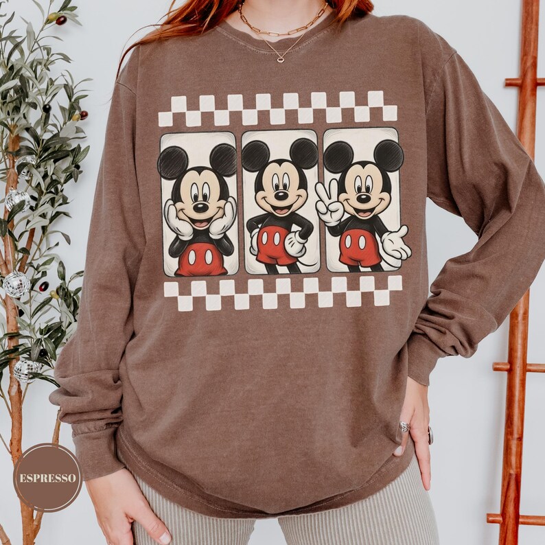 Puede incluir: Una camiseta marr&oacute;n de manga larga con un gr&aacute;fico de Mickey Mouse en tres poses diferentes. Las im&aacute;genes de Mickey Mouse est&aacute;n enmarcadas en cuadros blancos y negros. La camiseta tiene cuello redondo y un corte holgado.