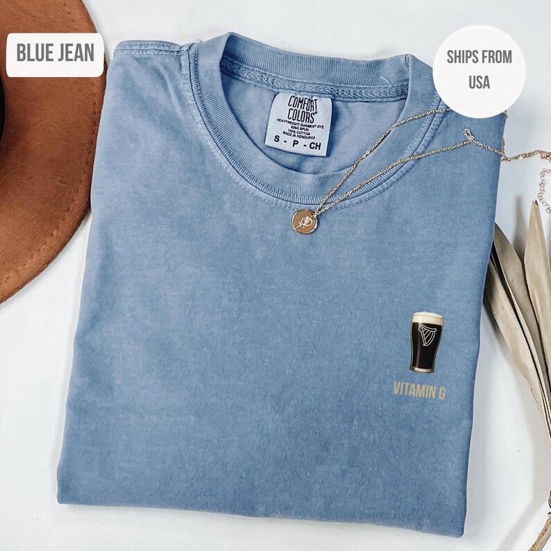May include: A blue jean colored Comfort Colors t-shirt with a gold necklace. The shirt features a graphic of a pint of stout with the text "Vitamin G". The shirt is a size small.