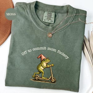 May include: Moss green t-shirt featuring the text "Off to commit mom foolery" and a frog on a scooter graphic. A gold necklace with a pendant rests on the shirt. The shirt is a Comfort Colors brand.