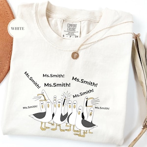 May include: White t-shirt with a graphic of seagulls yelling "Ms. Smith!" in black text. The shirt has a crew neck and short sleeves. A gold necklace with a pendant is draped across the shirt.