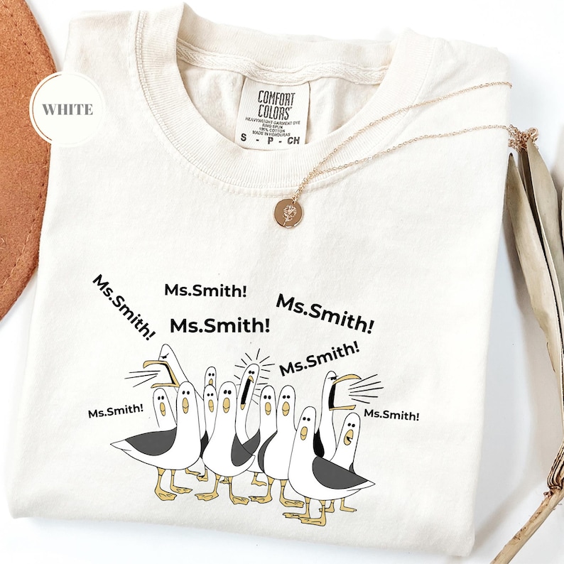 May include: White t-shirt with a graphic of seagulls yelling "Ms. Smith!" in black text. The shirt has a crew neck and short sleeves. A gold necklace with a pendant is draped across the shirt.