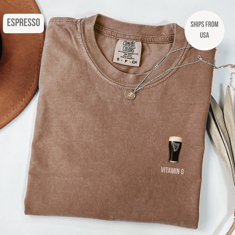 May include: Espresso-colored t-shirt with a crew neck and short sleeves. The shirt features a graphic of a pint of stout beer with the text "Vitamin G" below it. A silver necklace with a small pendant is draped across the shirt.