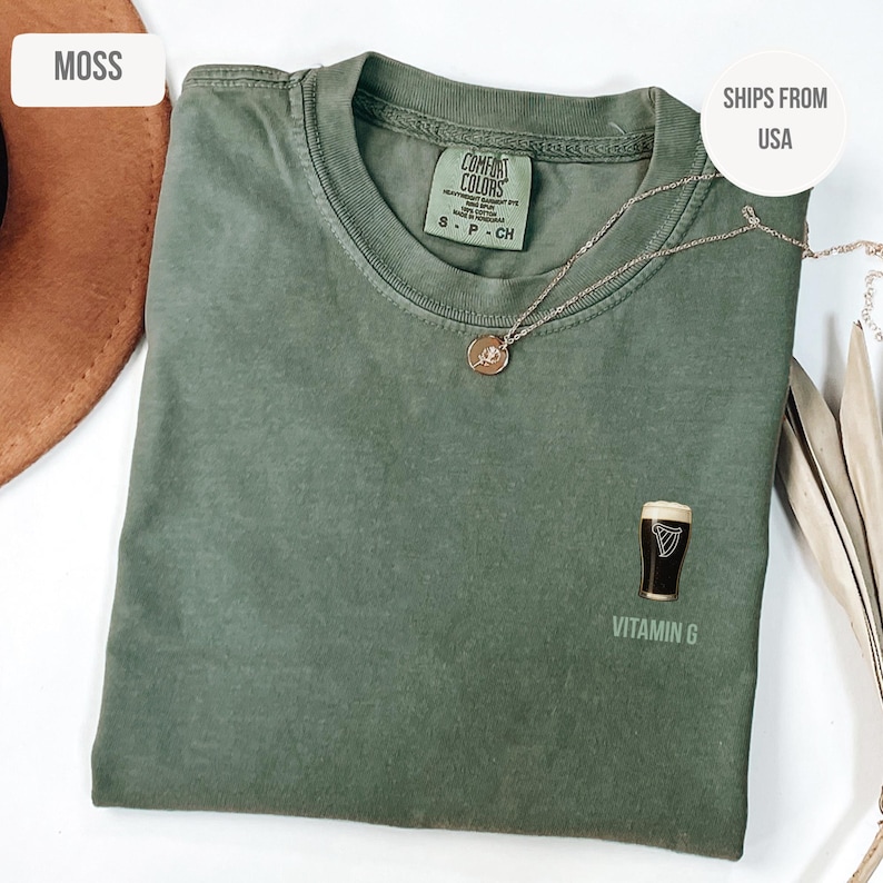 May include: A moss green t-shirt with a graphic of a pint of stout and the text "Vitamin G". The shirt has a crew neck and short sleeves. A silver necklace with a small pendant is draped across the shirt.