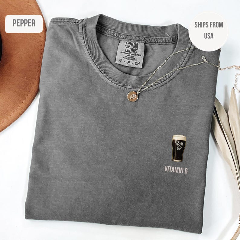 May include: A gray Comfort Colors t-shirt with a small graphic of a pint of stout and the text "Vitamin G." A silver necklace with a pendant is draped across the shirt.