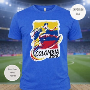 May include: A heather true royal blue t-shirt featuring a graphic of a soccer player kicking a ball, with the colors of the Colombian flag. The word "COLOMBIA" is arched below the graphic, with "2026" below it.