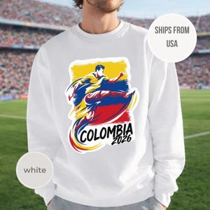 May include: White sweatshirt featuring a graphic of a soccer player in the colors of the Colombian flag. The design includes the word "COLOMBIA" and the year "2026". The sweatshirt is a crew neck style.