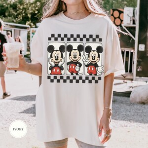 Puede incluir: Camiseta de color marfil con un gr&aacute;fico de Mickey Mouse en tres poses diferentes, sobre un patr&oacute;n de cuadros blancos y negros. Mickey lleva sus pantalones cortos rojos y guantes blancos. La camiseta tiene un corte holgado.