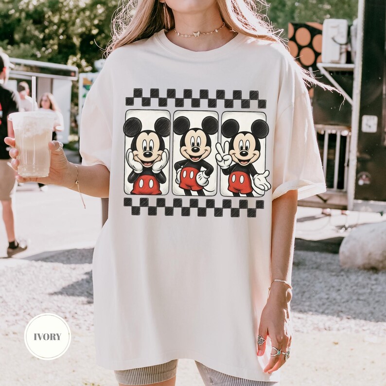 Puede incluir: Camiseta de color marfil con un gr&aacute;fico de Mickey Mouse en tres poses diferentes, sobre un patr&oacute;n de cuadros blancos y negros. Mickey lleva sus pantalones cortos rojos y guantes blancos. La camiseta tiene un corte holgado.