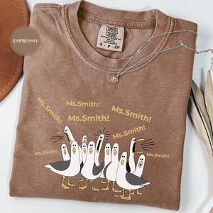 May include: Espresso-colored Comfort Colors t-shirt with a graphic of cartoon seagulls yelling "Ms. Smith!". The shirt has a crew neck and short sleeves. A silver necklace with a small pendant is draped across the shirt.