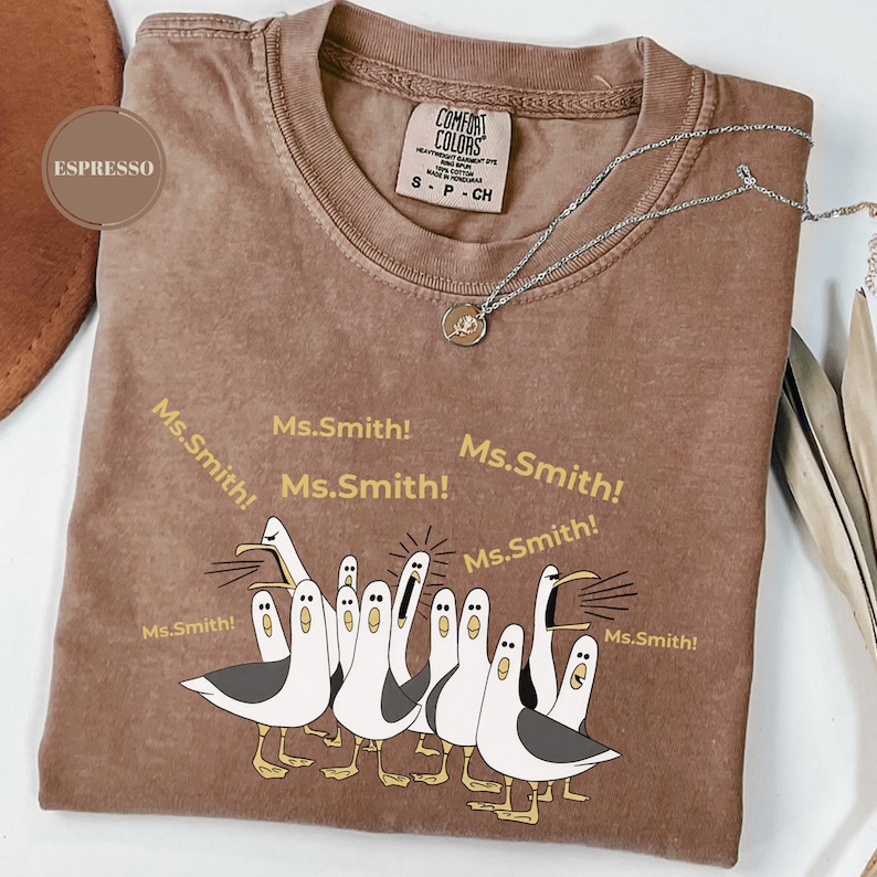 May include: Espresso-colored Comfort Colors t-shirt with a graphic of cartoon seagulls yelling "Ms. Smith!". The shirt has a crew neck and short sleeves. A silver necklace with a small pendant is draped across the shirt.