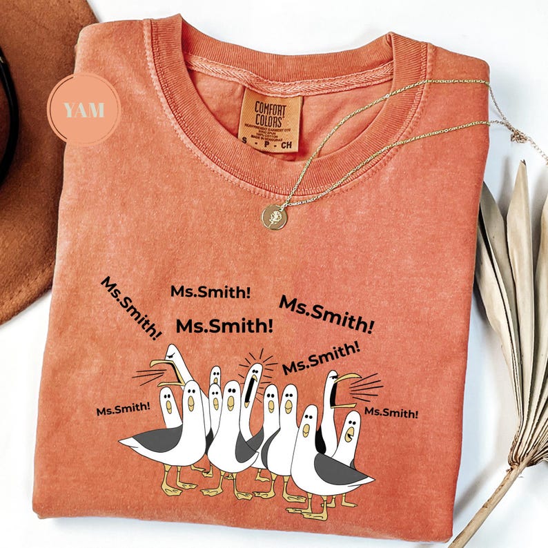 May include: Coral-colored Comfort Colors t-shirt featuring a cartoon illustration of seagulls with the text "Ms. Smith!" around them. The shirt has a crew neck and short sleeves. A gold necklace with a pendant is draped on the shirt.