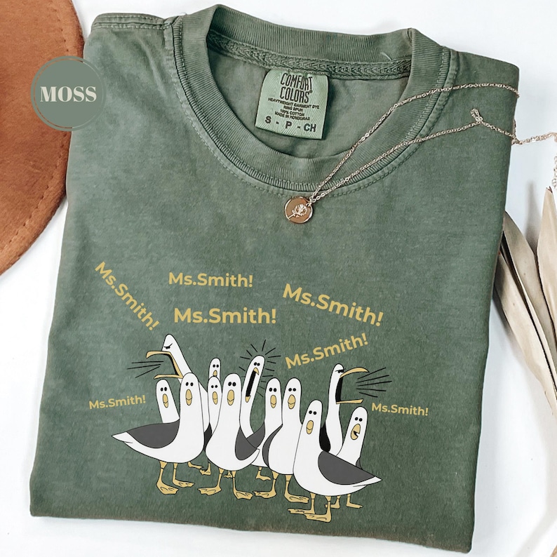 May include: Moss green Comfort Colors t-shirt with a graphic of cartoon seagulls yelling "Ms. Smith!". The shirt has a crew neck and short sleeves. A gold-colored necklace with a pendant is draped on the shirt.