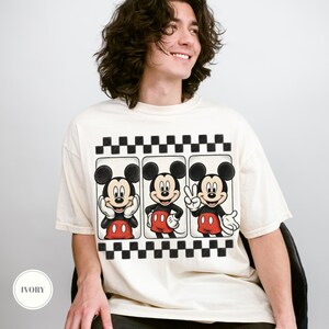 Puede incluir: Camiseta de color marfil con un dise&ntilde;o de Mickey Mouse en tres poses diferentes, con pantalones cortos rojos y guantes blancos. El dise&ntilde;o est&aacute; enmarcado por un borde a cuadros blanco y negro. La camiseta tiene un corte holgado.