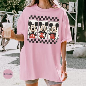 Puede incluir: Camiseta rosa claro de gran tama&ntilde;o con un gr&aacute;fico de Mickey Mouse en tres poses diferentes. Cada Mickey Mouse est&aacute; enmarcado por un borde a cuadros blanco y negro. La camiseta es de estilo casual y c&oacute;modo.