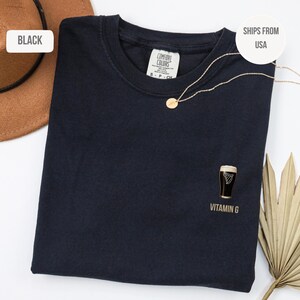 May include: Black t-shirt with a small graphic of a pint of stout and the text "Vitamin G". A gold necklace with a round pendant is draped across the shirt. A brown hat is visible in the upper left corner.