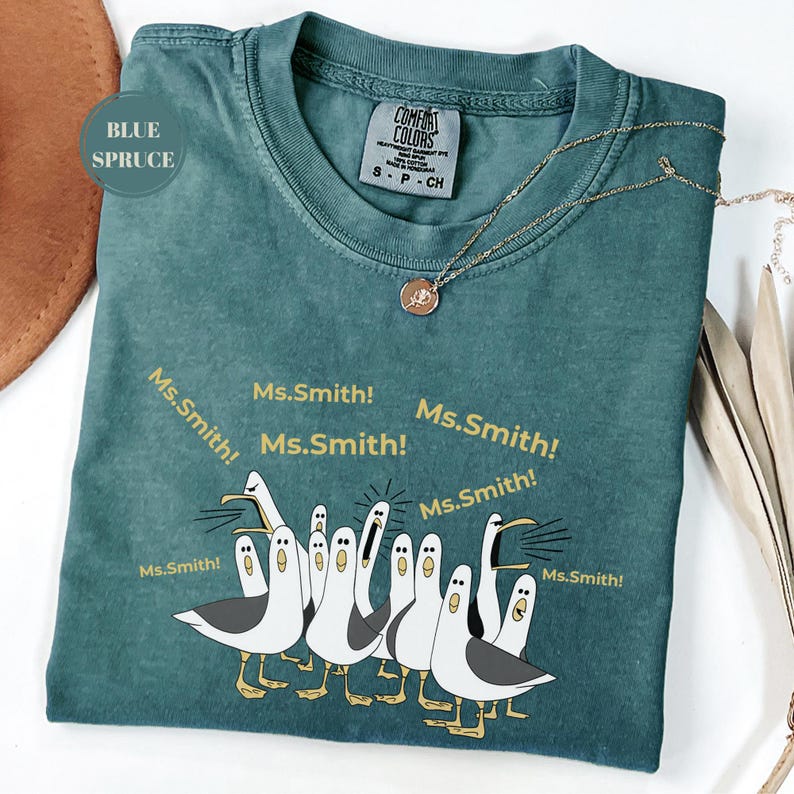 May include: Blue Spruce Comfort Colors t-shirt with a graphic of seagulls and the text "Ms.Smith!". A gold necklace with a pendant rests on the shirt. The shirt is size S-P-CH.