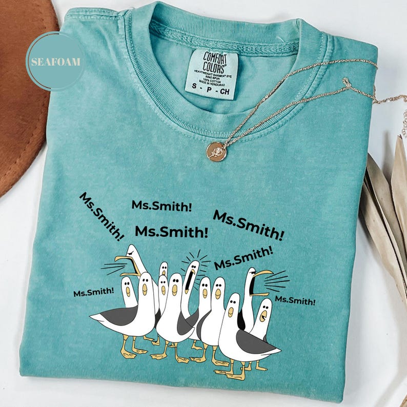 May include: Seafoam green Comfort Colors t-shirt with a graphic of seagulls yelling "Ms. Smith!". The shirt has a crew neck and short sleeves. A gold necklace with a pendant is draped across the shirt.