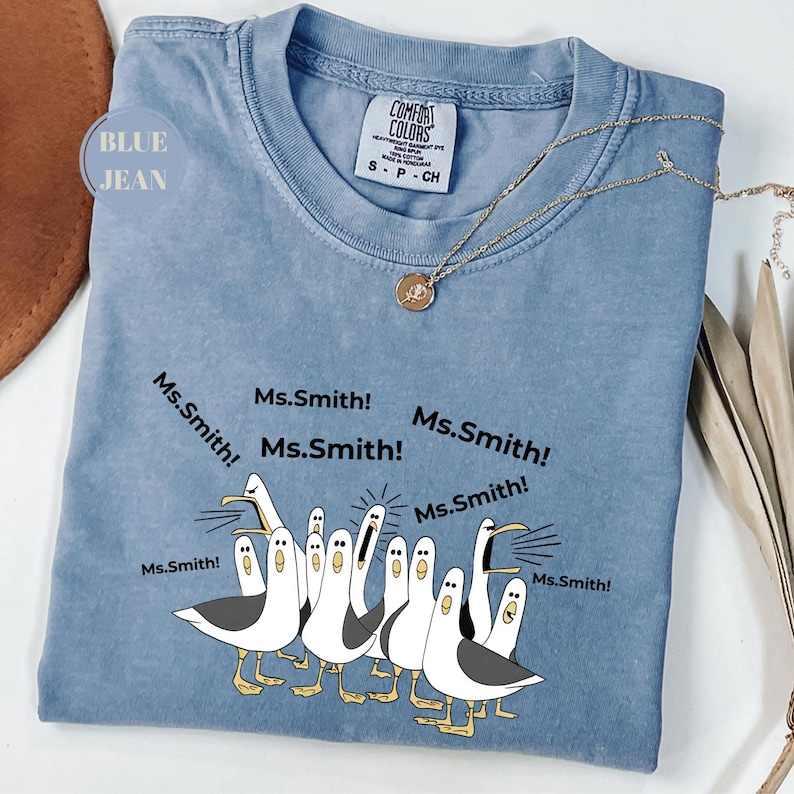 May include: A blue Comfort Colors t-shirt with a graphic of seagulls yelling "Ms. Smith!". The shirt has a crew neck and short sleeves. A gold necklace with a pendant is draped on the shirt.
