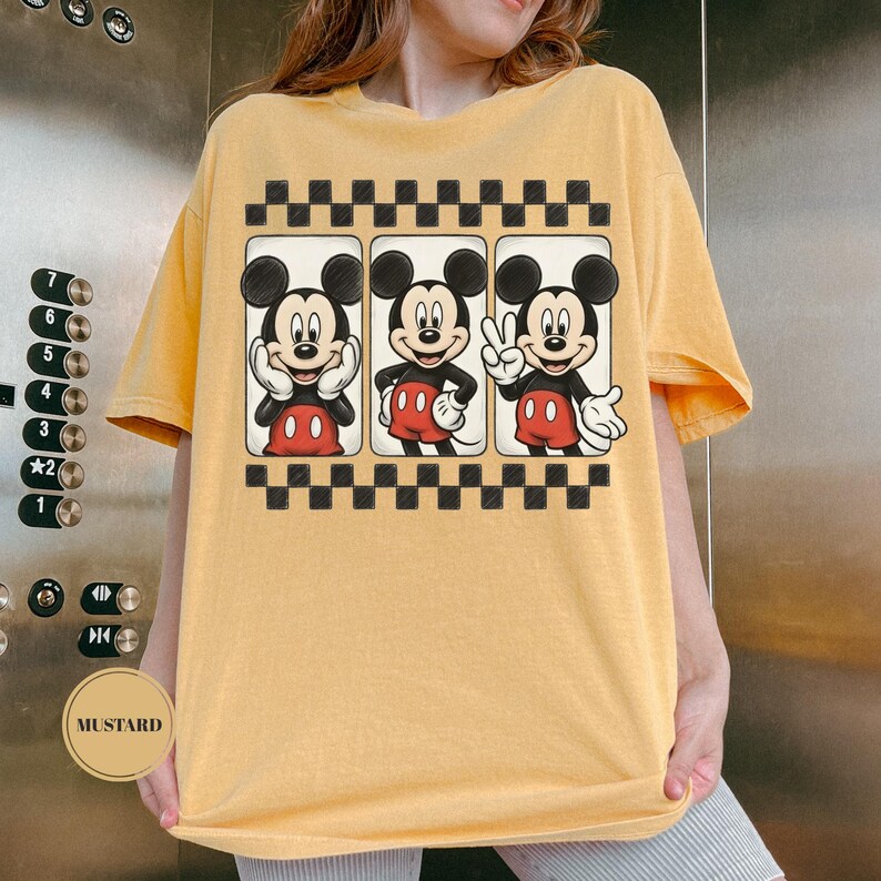 Puede incluir: Camiseta de color mostaza con un gr&aacute;fico de Mickey Mouse en tres poses diferentes. Mickey lleva sus cl&aacute;sicos pantalones cortos rojos y zapatos negros. El gr&aacute;fico est&aacute; enmarcado por un patr&oacute;n de cuadros blancos y negros.