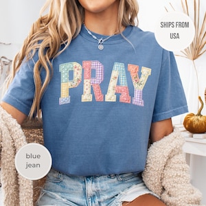 Puede incluir: Camiseta azul vaquero con la palabra "PRAY" en letras de patchwork. Las letras están en varios estampados florales y de cuadros vichy en tonos pastel. La camiseta tiene mangas cortas y cuello redondo.