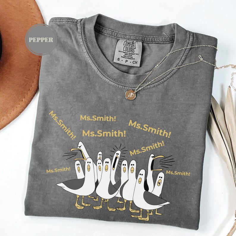 May include: A gray Comfort Colors t-shirt with a graphic of cartoon seagulls and the text "Ms. Smith!". The shirt has a crew neck and short sleeves. A silver necklace with a pendant is draped on the shirt.