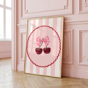 May include: A rectangular wall art print in a gold frame featuring a vertical light pink and white striped background. In the center, a scalloped red circular border frames two dark red cherries connected by a stem with a pink bow on top.