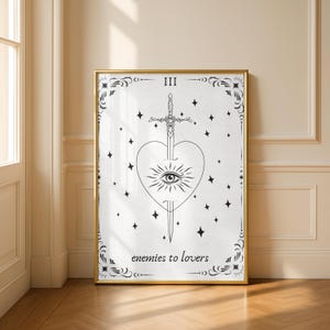 May include: A framed black and white tarot card print. The artwork features a heart pierced by a sword, an eye, and stars. The text "III" is at the top, and "enemies to lovers" is at the bottom. The frame is gold.