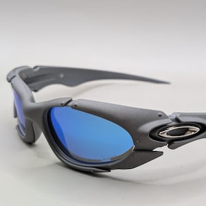 K&ouml;nnte beinhalten: Graue Sonnenbrille mit blauen Gl&auml;sern. Die Sonnenbrille hat ein sportliches Design mit matter Oberfl&auml;che. Die Gl&auml;ser sind leuchtend blau und die Rahmen sind ein ged&auml;mpftes Grau. Die B&uuml;gel der Sonnenbrille sind lang und schlank.