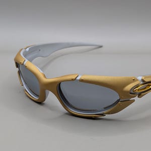 May include: Gold and white sunglasses with gray lenses. The sunglasses have a gold frame around the lenses and gold accents on the temples. The arms are white. The lenses are a dark gray color.