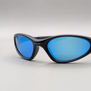 K&ouml;nnte beinhalten: Schwarze Sonnenbrille mit blau verspiegelten Gl&auml;sern. Die Sonnenbrille hat einen geschwungenen Rahmen und das Oakley-Logo ist auf der Seite sichtbar. Die Gl&auml;ser reflektieren das Licht und erzeugen ein hellblaues Aussehen.