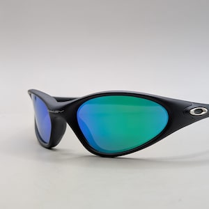 May include: Black sunglasses with a sleek, curved design. The lenses have a vibrant green and blue gradient. The frame features the Oakley logo on the temple. These sunglasses are designed for sports and outdoor activities.