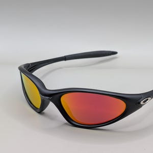 Pode incluir: &Oacute;culos de sol pretos com arma&ccedil;&atilde;o curva e lentes vermelhas e amarelas. Os &oacute;culos de sol t&ecirc;m um design elegante e moderno, com um pequeno logotipo prateado na lateral da arma&ccedil;&atilde;o. As lentes fazem uma transi&ccedil;&atilde;o do amarelo para o vermelho.