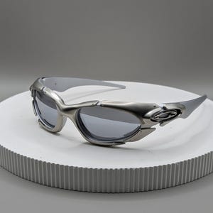 May include: Silver-framed sunglasses with gray lenses. The sunglasses have a futuristic design with a curved frame and a sleek, modern aesthetic. The lenses are a dark gray color, providing a contrast to the silver frame.