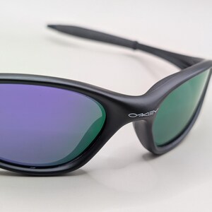 May include: Close-up of black sunglasses with purple and green lenses. The sunglasses have a sleek, modern design with the brand name "Oakley" printed on the side. The lenses reflect a gradient of colors.
