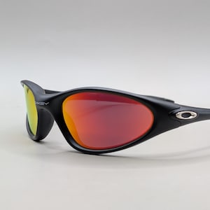 Pode incluir: &Oacute;culos de sol pretos com lentes vermelho-alaranjadas. Os &oacute;culos t&ecirc;m uma arma&ccedil;&atilde;o curva e um pequeno emblema prateado nas hastes. As lentes apresentam um efeito degrad&ecirc;, passando do vermelho para o laranja. Design desportivo.